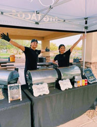 Two smiling servers with arms outstretched behind a black-draped outdoor catering table under a canopy, stainless-steel chafing dishes for a taco station, handwritten menu signs and canned drinks at a park pavilion
