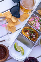 Top-down view of Mexican tacos in a metal tray—shredded meat tacos topped with chopped onion, cilantro and pickled red onion—served with tortilla chips, lime wedges, salsa and a beer on a white outdoor table