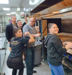 Smiling five-person kitchen crew cooking together in a commercial restaurant kitchen, woman at a flat‑top griddle and man pulling a tray from an oven under a stainless hood.