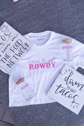 Flat-lay of a white t-shirt printed 'down to get ROWDY' in pink, two glass jars with wooden lids and matching pink lettering, and two rustic wood signs reading 'down to get tacos' and 'THE FOOD NETWORK' on a gray felt background — playful party-themed product shot.