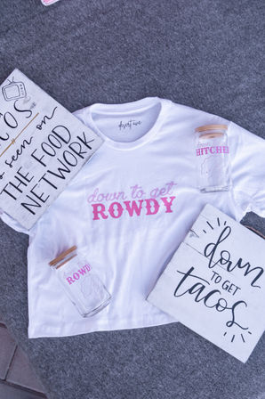Flat-lay of a white t-shirt printed 'down to get ROWDY' in pink, two glass jars with wooden lids and matching pink lettering, and two rustic wood signs reading 'down to get tacos' and 'THE FOOD NETWORK' on a gray felt background — playful party-themed product shot.