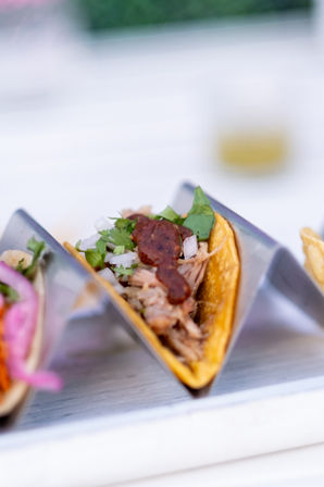 Juicy pulled-pork taco on a yellow corn tortilla in a metal taco holder, topped with salsa roja, diced white onion and cilantro on a white table.