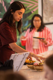 Woman serving food at an indoor buffet station, scooping from small brass bowls onto a plate; guest in a striped shirt holds a red cup with a tropical leaf mural backdrop.