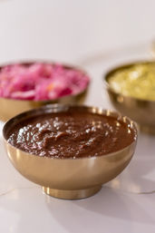 Close-up of brown chutney in a small brass bowl on a white tabletop, with blurred pink pickled onions and green chutney bowls in the background — colorful Indian condiments.
