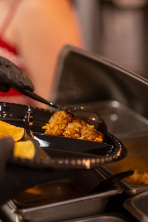 Steaming seasoned Mexican-style rice being scooped onto a black divided plastic plate with tortilla chips, served with a gloved hand and spoon at a buffet station.