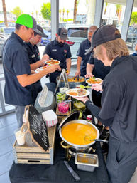 Team in black uniforms serving themselves at an indoor buffet in an auto showroom — large pot of creamy orange soup, tacos, salsas, pickled onions and lime wedges.