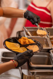 Gloved server scoops creamy refried beans onto a divided black plate with corn tortillas and Mexican rice at a buffet-style catering station