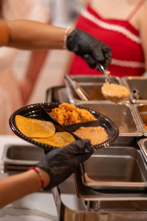 Gloved server scoops creamy refried beans onto a divided black plate with corn tortillas and Mexican rice at a buffet-style catering station