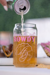 Golden beer poured from a can into a clear taco-themed glass with pink lettering, tacos and a tortilla chip blurred in the outdoor food scene