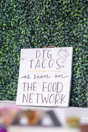 Hand-painted wooden sign advertising tacos with “as seen on The Food Network” text, propped against a green boxwood wall at an outdoor food stand