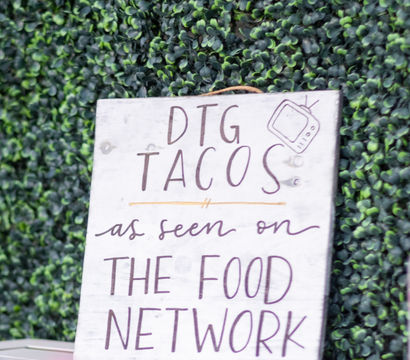Hand-painted wooden sign advertising tacos with “as seen on The Food Network” text, propped against a green boxwood wall at an outdoor food stand