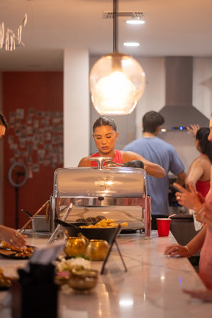 Person serving food from a chafing dish on a marble kitchen island at a casual indoor buffet party beneath a warm pendant light.