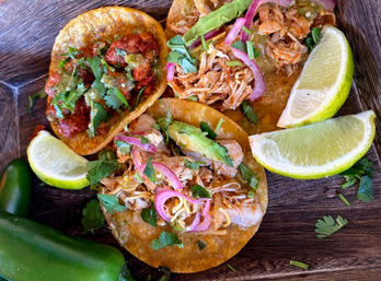 Three Mexican-style tacos on corn tortillas topped with shredded chicken, pickled red onions, avocado, cilantro and salsa, served with lime wedges and jalapeños on a wooden board.