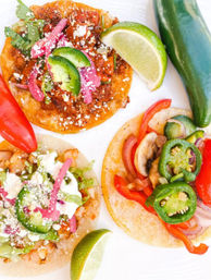 Three colorful street-style Mexican tacos on corn tortillas topped with shredded meat and sautéed vegetables, pickled red onions, sliced jalapeños, crumbled queso and fresh lime wedges
