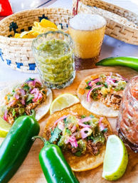 Three Mexican street tacos on corn tortillas topped with shredded meat, salsa verde, pickled red onions and cilantro on a wooden board with lime wedges, fresh jalapeños, a basket of tortilla chips and a frothy amber beer.