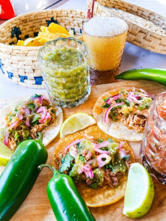 Three Mexican street tacos on corn tortillas topped with shredded meat, salsa verde, pickled red onions and cilantro on a wooden board with lime wedges, fresh jalapeños, a basket of tortilla chips and a frothy amber beer.
