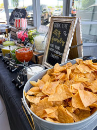 Metal tub of crispy tortilla chips at a self-serve nacho bar with bowls of salsa, guacamole and sour cream and a chalkboard sign in a bright indoor catering setup