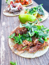Close-up of pulled pork street tacos on corn tortillas, topped with fresh chopped cilantro and a lime wedge on a rustic wooden board.