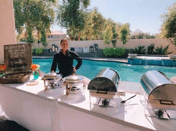 Sunny backyard poolside catering scene with white buffet table, stainless-steel chafing dishes, small floral arrangements, and a staff member by a turquoise swimming pool.