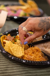 Close-up of a hand with blue nail polish and rings snagging a tortilla chip from a black plastic plate of Mexican food with rice and tacos.