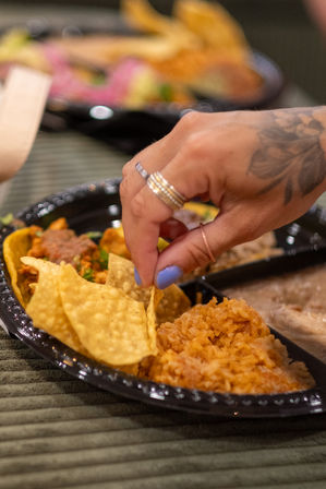 Close-up of a hand with blue nail polish and rings snagging a tortilla chip from a black plastic plate of Mexican food with rice and tacos.