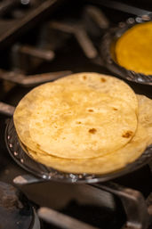 Stack of warm corn tortillas on a glass plate resting on a gas stovetop burner, golden-browned spots, ready for tacos