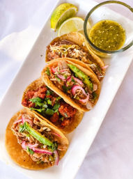 Four Mexican street-style tacos on a white rectangular plate — shredded pork, chorizo, pickled red onions and avocado, served with lime wedges and a small glass of green salsa