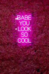 Bright pink neon sign reading 'BABE YOU LOOK SO COOL' mounted on a dense pink rose flower wall — trendy Instagram-ready floral photo backdrop.