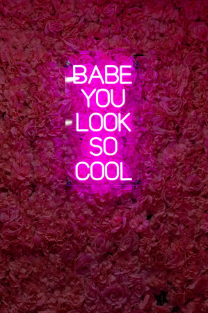 Bright pink neon sign reading 'BABE YOU LOOK SO COOL' mounted on a dense pink rose flower wall — trendy Instagram-ready floral photo backdrop.