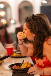 Woman with sunglasses on her head mid-bite into a taco above a plate of tortilla chips and toppings at a lively indoor eatery.