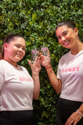 Two smiling women in white bachelorette shirts with pink lettering clinking clear glasses in front of a green foliage wall backdrop, festive indoor celebration photo.