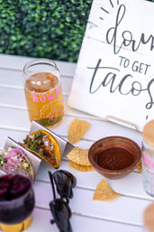 Outdoor patio taco spread with two street tacos in metal holders topped with cilantro and onions, tortilla chips, a wooden bowl of red salsa, craft beer glass and a "down to get tacos" sign.