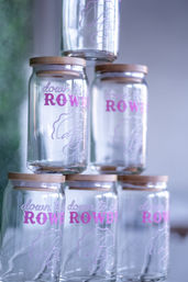 Stacked clear glass jars with bamboo lids and bright pink 'ROWDY' lettering, arranged in a playful pyramid display on a soft, blurred background.