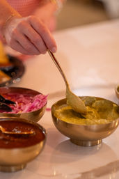 Hand with gold spoon scooping green chutney from a brass bowl beside pickled red onions and spicy chutney on an Indian restaurant table
