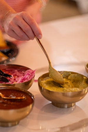 Hand with gold spoon scooping green chutney from a brass bowl beside pickled red onions and spicy chutney on an Indian restaurant table