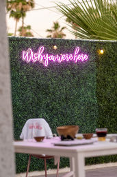 Pink neon 'Wish you were here' sign on a lush green hedge wall with string lights, palm trees in the background and a casual outdoor patio table with bowls and a drink.
