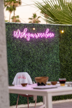 Pink neon 'Wish you were here' sign on a lush green hedge wall with string lights, palm trees in the background and a casual outdoor patio table with bowls and a drink.