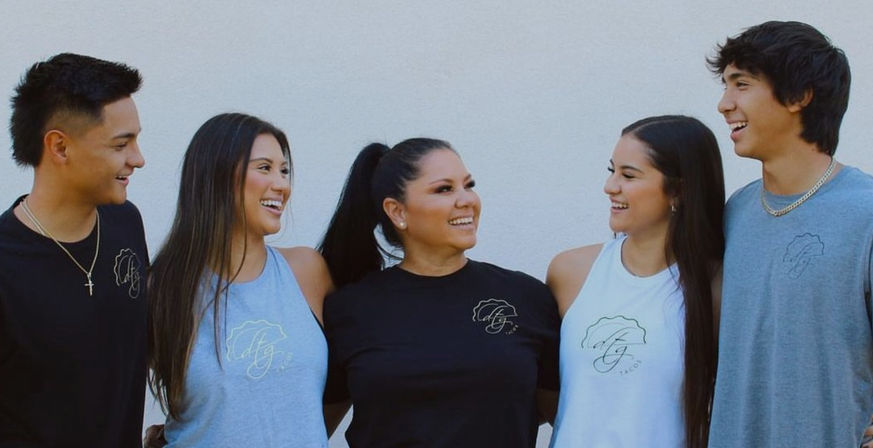 Five smiling people in casual branded tees and tank tops stand against a white wall, sharing laughs in a lifestyle apparel group portrait.