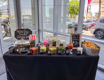 Nacho bar in a bright glass-walled showroom with a metal tub of tortilla chips, chafing dish and bowls of red and green salsas, guacamole, jalapeños, cheese and sour cream, labeled with small chalkboard signs on a black tablecloth.