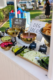 Outdoor backyard taco bar with golden bowls of salsa and guacamole, tortilla chips, lime wedges, chopped onions, cilantro and pickled red onions, plus chalkboard signs — bright fiesta-style toppings spread for a sunny garden party.
