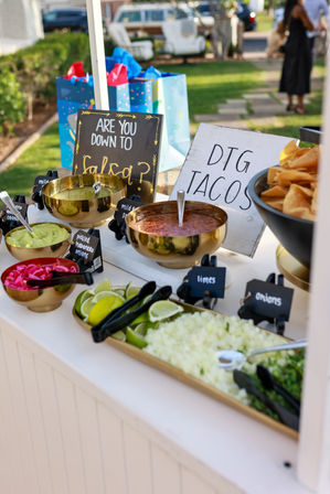 Outdoor backyard taco bar with golden bowls of salsa and guacamole, tortilla chips, lime wedges, chopped onions, cilantro and pickled red onions, plus chalkboard signs — bright fiesta-style toppings spread for a sunny garden party.