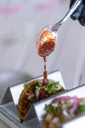 Close-up of a spoon drizzling spicy red salsa onto a shredded-meat Mexican street taco topped with chopped onion and cilantro in a metal taco holder