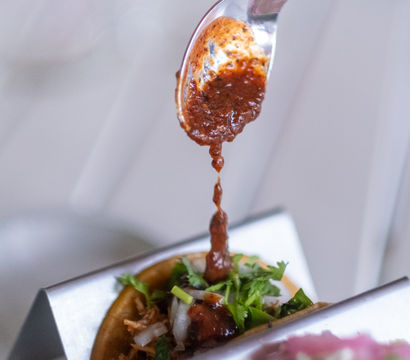 Close-up of a spoon drizzling spicy red salsa onto a shredded-meat Mexican street taco topped with chopped onion and cilantro in a metal taco holder
