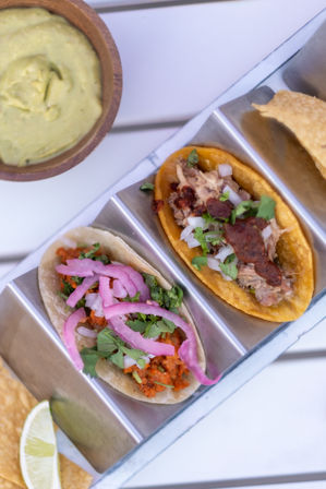 Top-down close-up of two Mexican street tacos in a stainless taco holder — one corn tortilla with shredded meat, diced onion, cilantro and red salsa; the other with seasoned meat, pickled red onions and cilantro, plus a bowl of guacamole, lime wedge and tortilla chips on a white table
