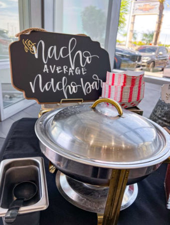 Playful “Nacho Average Nacho Bar” chalkboard sign behind a covered stainless chafing dish with ladle and red-striped paper trays on a catered buffet table by a window with parked cars outside.