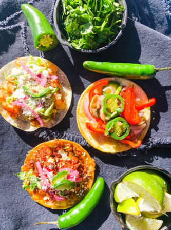 Overhead shot of three vibrant Mexican street tacos on corn tortillas topped with pickled onions, crumbled cheese, sliced jalapeño, avocado and sautéed peppers, with whole jalapeños, a bowl of chopped cilantro and lime wedges on a dark cloth