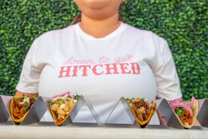 Row of fresh street tacos in a metal rack on a counter, with a person wearing a white t-shirt that reads 'down to get HITCHED' and a green hedge backdrop