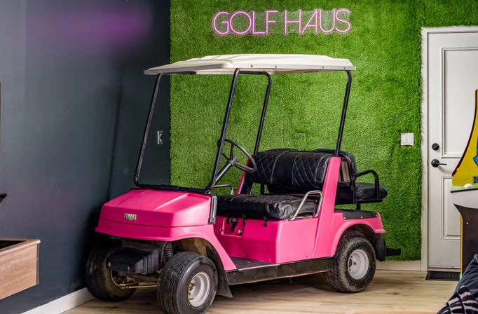 Bright pink golf cart parked indoors against a green artificial-turf wall with a neon golf sign, black leather seats and light wood flooring — trendy indoor golf lounge vibe.