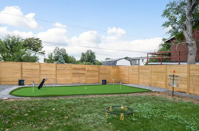 Backyard with a synthetic putting green, golf bag and practice flags, wooden privacy fence, disc golf practice basket with red flag, and a Spikeball set on the lawn under a blue sky.