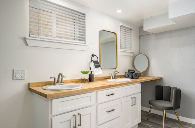 Bright modern bathroom with double-sink white vanity, wood countertop, gold-framed mirrors, rolled towels and a gray accent chair.